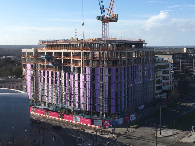 The Moxy Hotel in the construction phase, shows the complete square building from a distance with scaffold and purple building wrap