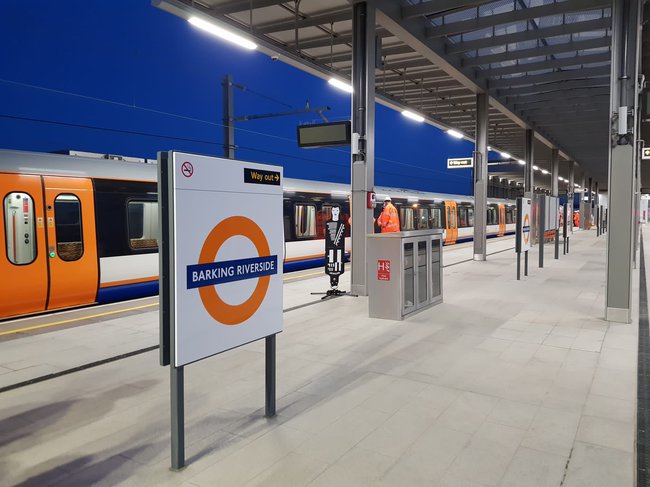 A train sits at a platform. London Overground signage for Barking Riverside is visible in the foreground