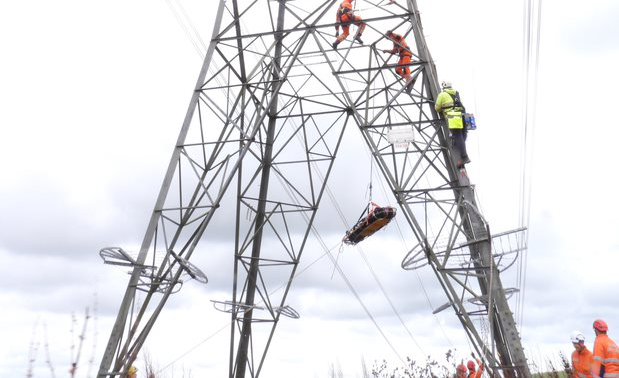A photograph showing a large electricity pylon. Three people wearing Personal Protective Equipment (PPE) are visible climbing up the tower as part of an exercise. A stretcher is being lowered from the pylon.