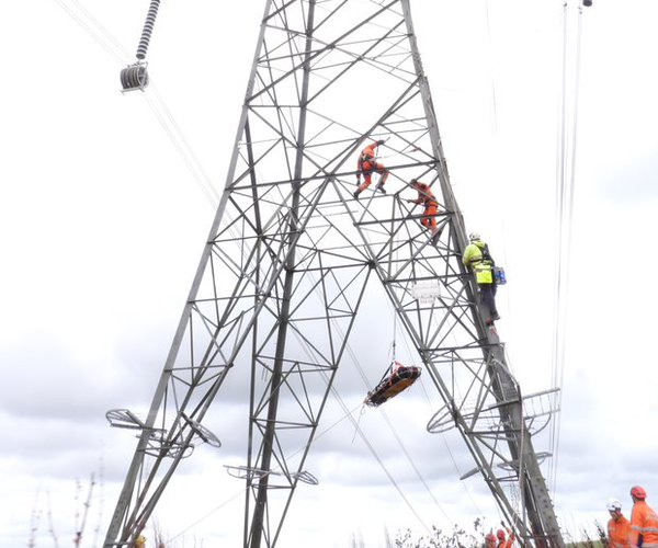 A photograph showing a large electricity pylon. Three people wearing Personal Protective Equipment (PPE) are visible climbing up the tower as part of an exercise. A stretcher is being lowered from the pylon.