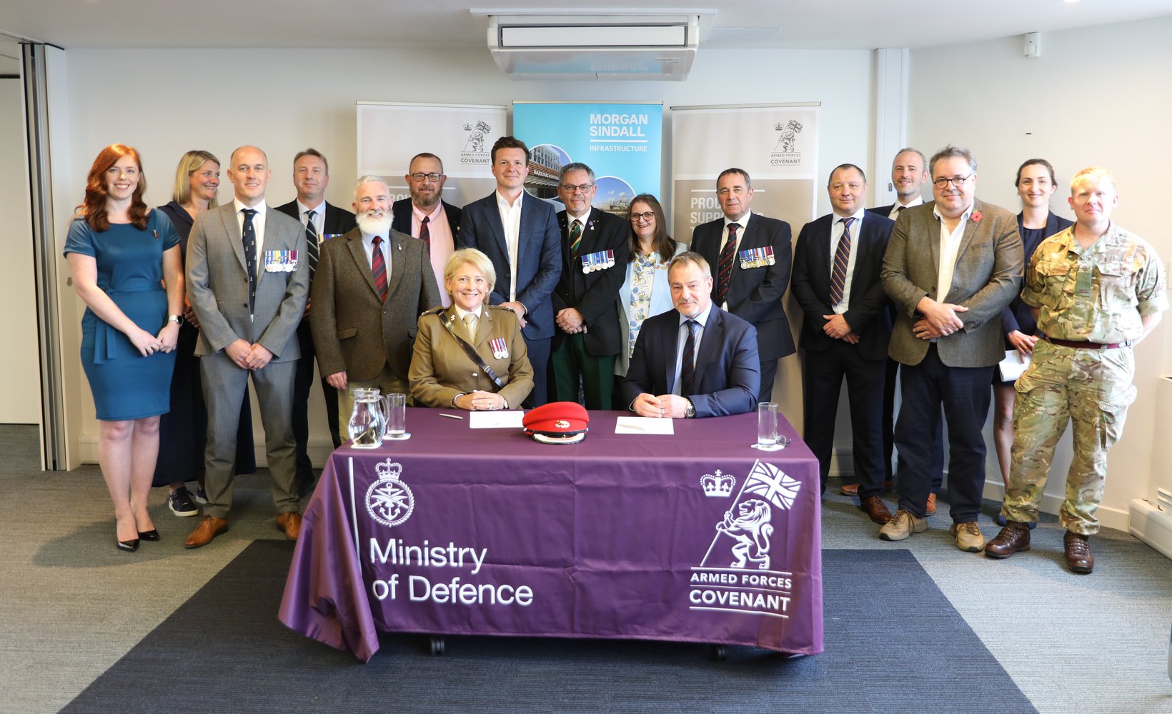 Major S Brettell DL of the Ministry of Defence, and Simon Smith, managing director of Morgan Sindall Infrastructure, are pictured seated at a table, with several Morgan Sindall Infrastructure colleagues standing behind them