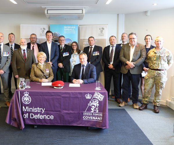 Major S Brettell DL of the Ministry of Defence, and Simon Smith, managing director of Morgan Sindall Infrastructure, are pictured seated at a table, with several Morgan Sindall Infrastructure colleagues standing behind them