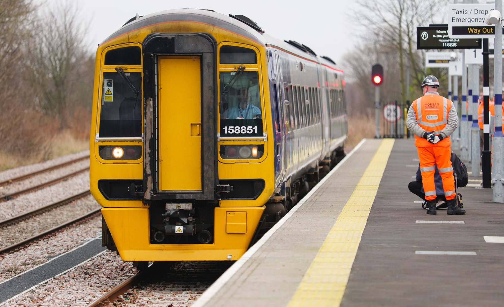A yellow and blue train stops at a platform. A worker in an orange vest stands nearby, with a red signal light in the background.