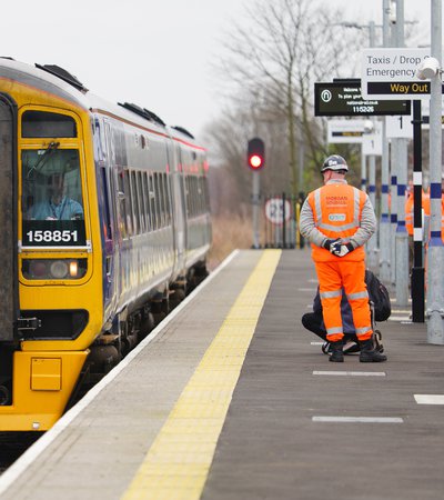 A yellow and black train is stopped at a platform. A worker in an orange safety suit and helmet stands nearby with their hands behind their back.