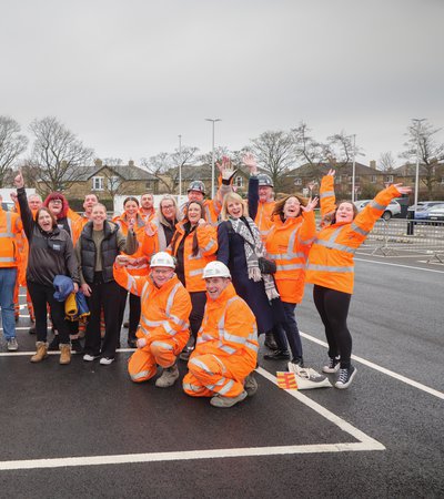 A group of workers in orange vests and others in casual clothes cheerfully pose together, raising their arms.