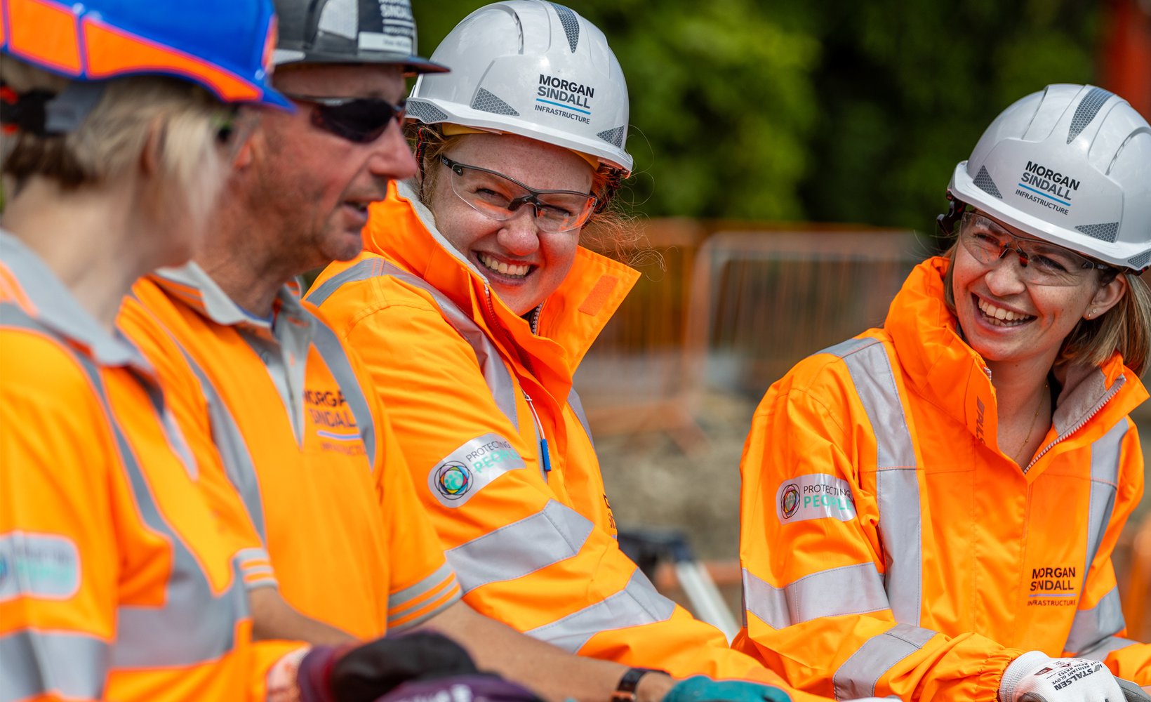 Four colleagues smile on site, standing at some temporary fencing in orange high visibility-wear.