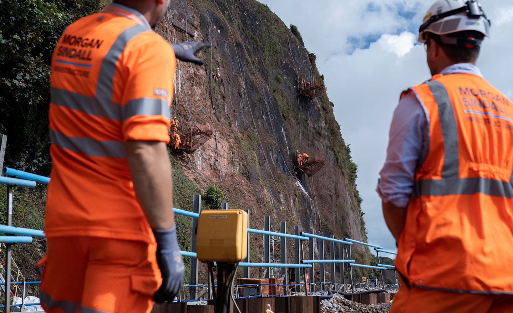 Two Morgan Sindall Infrastructure employees wearing Personal Protective Equipment (PPE) stand with their backs to the camera. They are facing the cliffside overlooking Parson's Tunnel, where some abseiling team members can be seen working on the rock