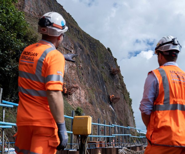 Two Morgan Sindall Infrastructure employees wearing Personal Protective Equipment (PPE) stand with their backs to the camera. They are facing the cliffside overlooking Parson's Tunnel, where some abseiling team members can be seen working on the rock
